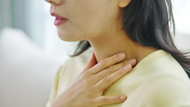 Close-up of an Asian woman touching her throat, indicating discomfort, sore throat, or a health issue like a cold or flu. Healthcare and wellness concept.