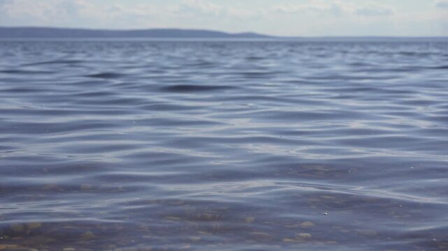 Calm, clear water creating gentle ripples and small waves on the surface, revealing a shallow, pebbly bottom with a distant, hazy shoreline under a bright, cloudy sky in the background