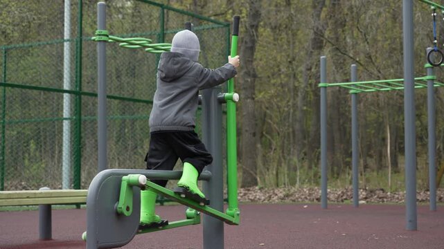 A spirited young child, dressed in a warm gray hoodie and striking green boots, enjoys an invigorating workout on a bright green outdoor elliptical machine.