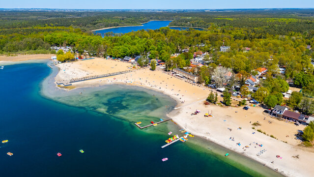 Wide aerial drone view of the holiday resort in Skorzecin, featuring a large sandy beach by Niedziegiel lake, wooden piers, holiday cabins, recreational infrastructure, pedal boats, surrounding green 
