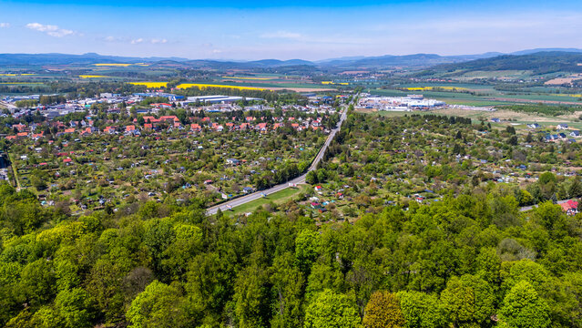 Wide aerial drone view of Klodzko town, featuring green allotment gardens, residential houses, a commercial area, and a road cutting through the landscape, with a lush forest in the foreground and mou