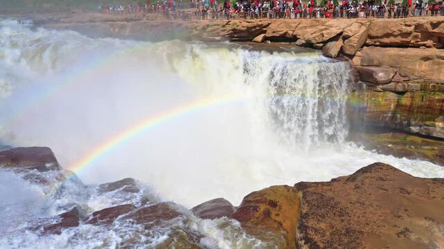 Detailed high-angle shot looking down at the crest of Hukou Waterfall. A vivid double rainbow stretches across the churning white water and rising mist as the Yellow River surges forward.