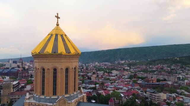 Detailed drone shot of the golden dome and upper architecture of the Holy Trinity Cathedral (Sameba) in Tbilisi, Georgia, with a panoramic view of the city and surrounding mountains in the background