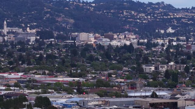 Aerial view of downtown Berkeley, California, with the Berkeley hills in the background, showing the campanile tower of UC Berkeley and the urban landscape with dense trees and buildings