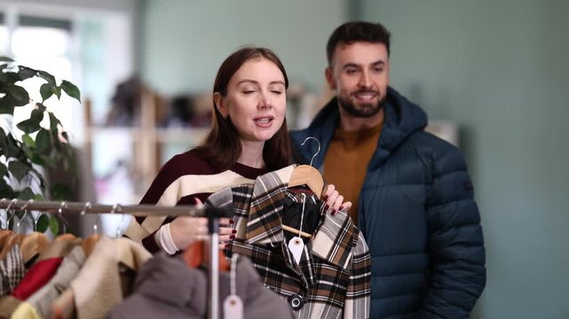 Woman holds a wool coat next to a man who tries on a dark blue down jacket. Buying women and men outerwear. High quality 4k footage