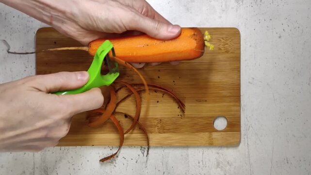 Hands peeling fresh orange carrot with green peeler on wooden board, healthy cooking preparation, kitchen vegetable processing, diet nutrition concept.