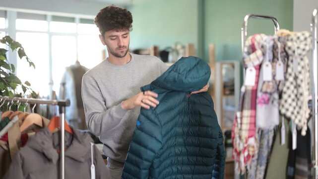 Fashionista guy client pre-examine fabric and style of product, is busy choosing down jacket. Client enjoys shopping for clothes at store browsing through colorful on display racks