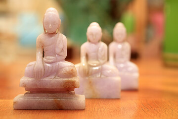 Three pale pink jade Buddha statues are arranged on a wooden surface. The first statue is in focus  the other two are blurry. © Godong Photo