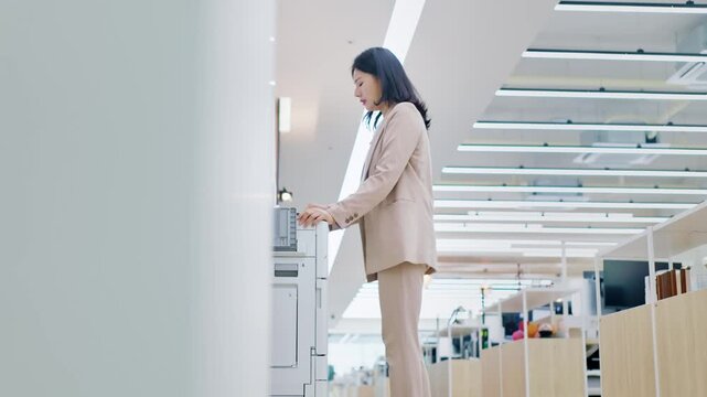 Focused Asian business woman operating a modern office printer or copier machine in a bright, contemporary workspace, highlighting efficiency and technology in a corporate environment.