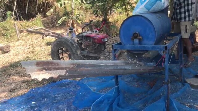 Rural Rice Processing: A farmer operates a simple mechanical threshing machine, separating rice grains in a sun-drenched field, showcasing traditional agricultural practices in a rural setting.
