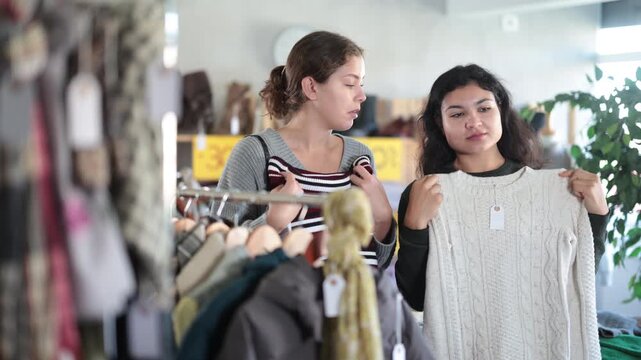 European woman and a Latin woman are standing in a store on background of shelves with clothes, they are talking and choosing a warm sweater. Two shoppers looking at jersey sweaters at discounts. High
