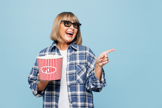 Elderly woman 50s years old wear shirt casual clothes 3d glasses watch movie film hold bucket of popcorn in cinema point finger aside on workspace area isolated on plain pastel light blue background.