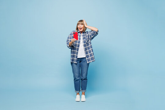 Full body shocked surprised elderly blonde woman she wear checkered shirt casual clothes hold in hand use mobile cell phone isolated on plain pastel blue background studio portrait. Lifestyle concept.