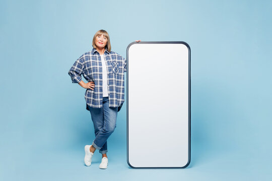 Full body side view elderly blonde woman 50s years old wearing checkered shirt casual clothes stand near big huge blank screen mobile cell phone smartphone with area isolated on plain blue background.