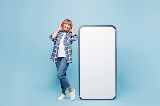 Full body elderly blonde woman 50s year old wear checkered shirt casual clothes big blank screen mobile cell phone smartphone with area listen music in headphones isolated on plain blue background