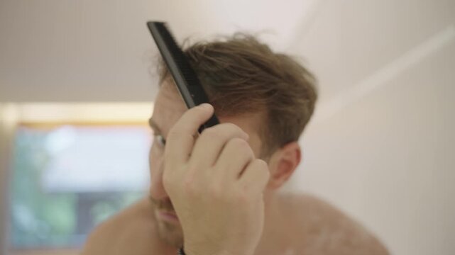 Young man with a beard looking in the mirror while styling his hair with a comb