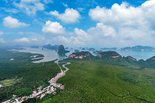 Limestone formations rise above emerald waters fringed by winding mangroves with villages and ports against a backdrop of cumulus clouds in a tropical landscape