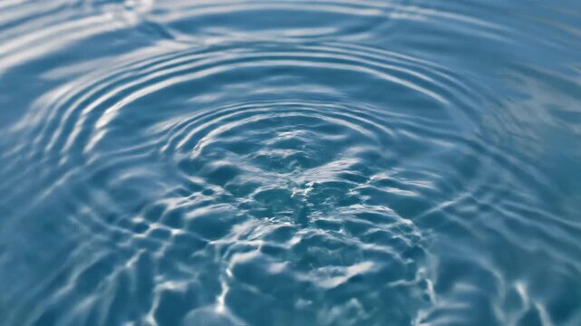 High-speed macro shot of a water droplet impacting a calm blue liquid surface, creating expanding concentric circular ripples and a vertical rebound splash with sky reflections.