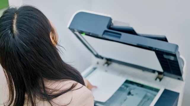 Woman operating a multi-function printer and copier machine in an office, placing a document on the scanner glass for copying or scanning.