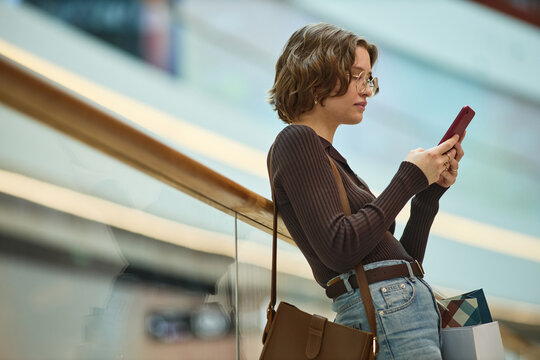 Young adult Caucasian woman checking smartphone while carrying shopping bags in mall, using mobile retail and messaging. Ideal for e commerce, consumer behavior, digital shopping campaigns