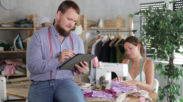 Young bearded tailor in checked shirt sitting on edge of worktable, sketching clothing designs on tablet with focused, creative look while female colleague working at sewing machine in cozy workshop