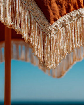 Close-up of cream macrame fringe detail on rust orange beach umbrella against blue sky, boho summer handcraft texture background