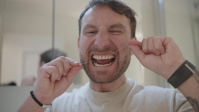 Humorous young man with a beard making exaggerated faces while flossing his teeth in the bathroom