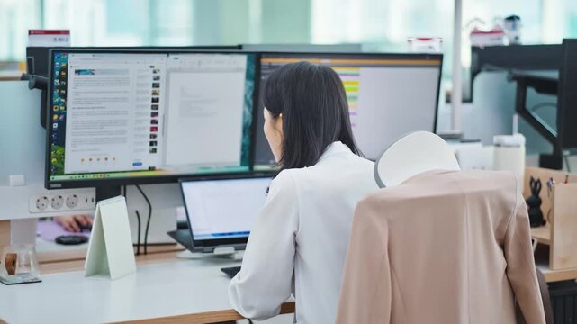 Dedicated Asian businesswoman focused on her work at a modern office desk with dual monitors and a laptop, showcasing productivity and a professional corporate environment.