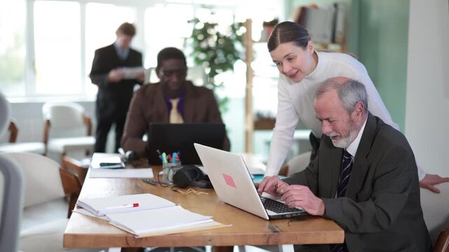 Woman leaned over to seated mature man employee, helps with work and looks at laptop screen. High quality 4k footage
