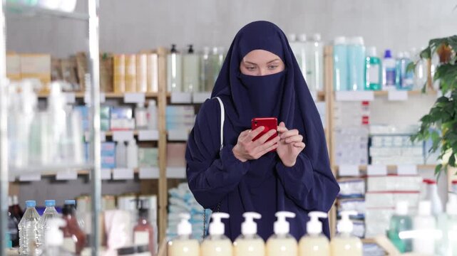 Young Muslim woman in traditional islamic navy blue dress and niqab covering face except eyes, shopping for medications or care products in drugstore, using phone to check list of needed items