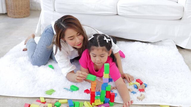 Asian mother and daughter playing colorful plastic blocks together on a white fluffy rug in a bright living room, fostering creativity and family bonding time at home.