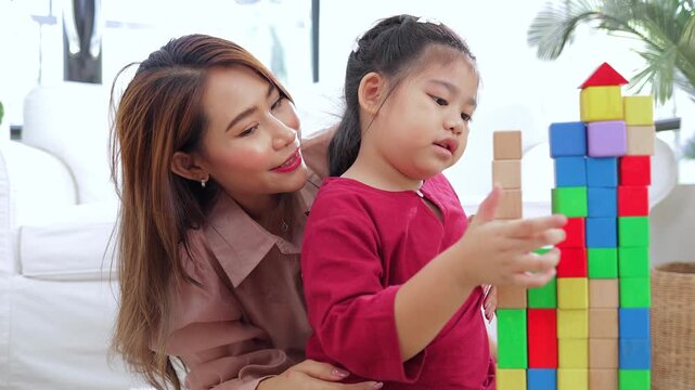Happy Asian girl concentrating on stacking colorful toy bricks with her mother nearby. Concept of early childhood development, fine motor skills, and home education. 