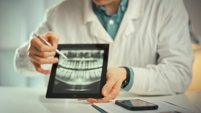 A tablet displaying a panoramic dental image sits in a dental office as a doctor in a white coat guides the patient through a consultation, highlighting problem teeth and outlining the treatment plan