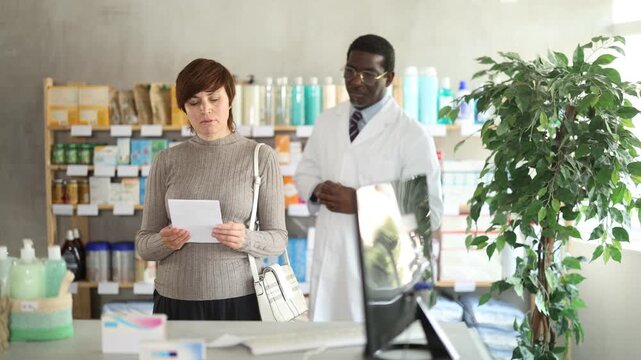In sales area of drugstore, woman with prescription looks at display cases, trying to find multivitamins for adults in open display. African male pharmacist informs about possibility of replacing