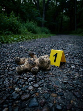 Abandoned teddy bear, mud-covered, next to crime scene marker on dark gravel path, representing loss or investigation.