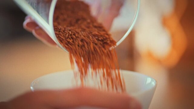 In home kitchen, housewife carefully pours brown rice from a storage jar into a white bowl, initiating the process of preparing rice grains for a nutritious meal, showcasing her daily cooking routine