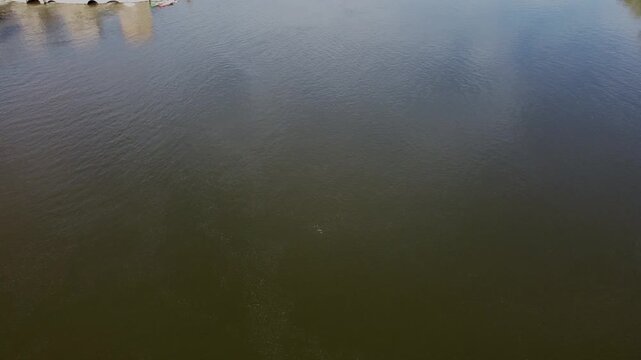 4K aerial drone video flying very low over the brown water of the Duero River in Zamora, Spain. The slow forward movement reveals a small island with a house and moored boats on a bright sunny day
