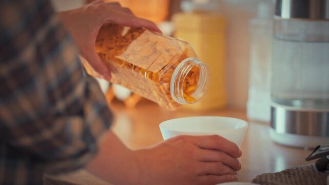 A woman's hands carefully pour yellow cornflakes into a white bowl, preparing a low-calorie morning breakfast at home, reflecting her commitment to conscious nutrition and a healthy lifestyle