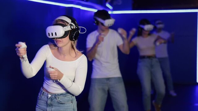 Girl in vr glasses with earbuds and two joysticks in hands is playing an active video game with friends. Female gamer in a virtual reality helmet plunged with friends into simulation