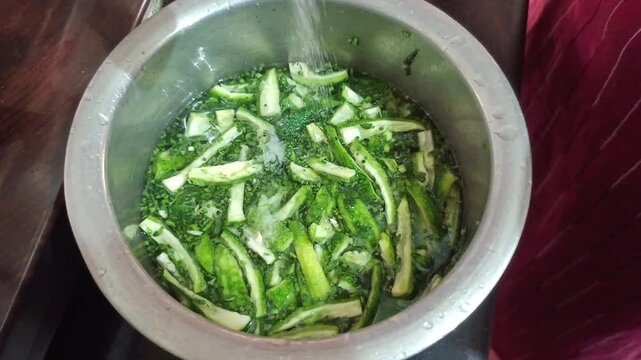 Adding white salt to chopped bitter gourd slices soaked in a steel bowl of water, reducing bitterness of karela