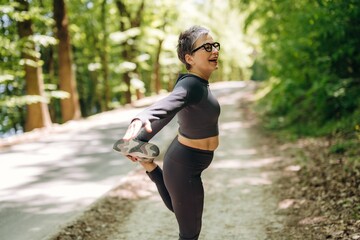 Fototapeta premium Middle-aged woman in black activewear stretches leg while smiling outdoors on a forest path surrounded by green trees and sunlight filtering through the leaves