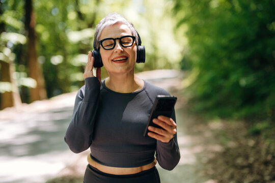 Mature woman with short gray hair wearing headphones listens to music on smartphone while walking along a forest path surrounded by greenery and sunlight