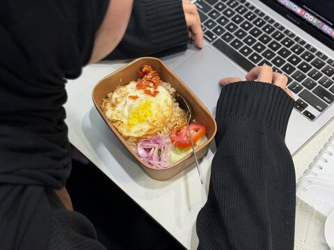 Hijab woman eating boxed fried rice while working in front of a laptop. Represents overtime culture, late working hours, and everyday hard work lifestyle in Indonesia.