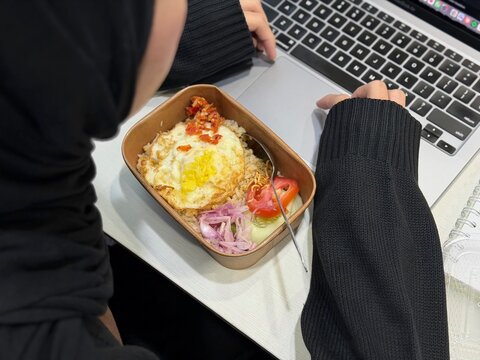Hijab woman eating boxed fried rice while working in front of a laptop. Represents overtime culture, late working hours, and everyday hard work lifestyle in Indonesia.