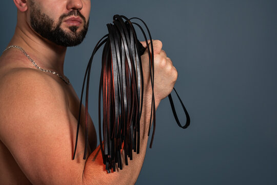 Studio close up of a shirtless bearded man holding a black leather flogger whip for adult BDSM and fetish concept