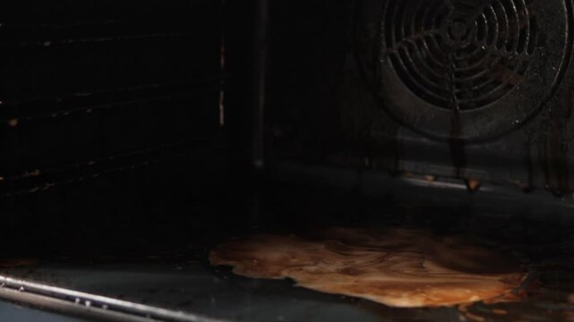 A person wipes the inner surface of a very dirty oven with a rag. A puddle of cleaning detergent and dissolved old grease has accumulated at the bottom
