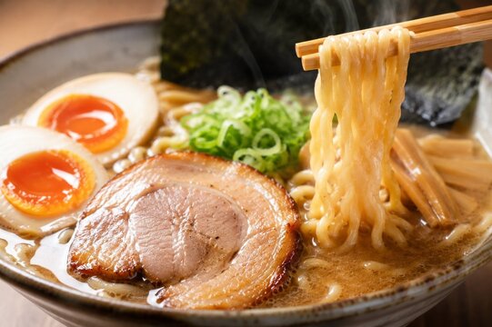 Authentic Japanese ramen bowl with chashu pork, soft-boiled egg, and fresh noodles lifted by chopsticks &mdash; rich tonkotsu broth, steam rising, dark ceramic bowl, close-up food photography