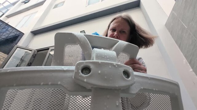 Washing mesh backrest of seat shows woman scrubbing white computer chair from low angle view. Drops of water fall down during washing mesh backrest of seat on outdoor patio.