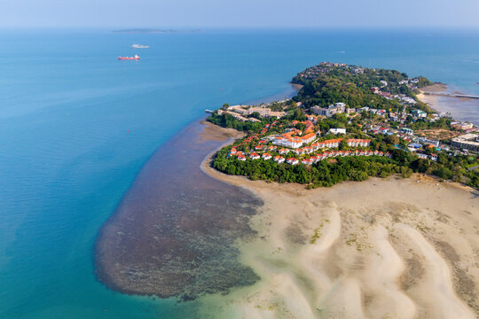 Coastal settlement with homes and lush trees borders wide sandy tidal flats merging into the deep blue ocean. Distant cargo ships sail under a clear sky
