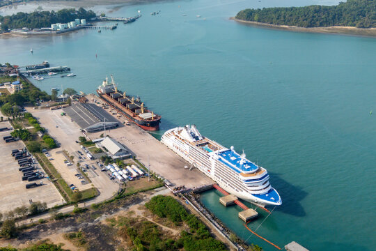 Docked at a vibrant port, a majestic cruise ship and a robust cargo vessel sit in clear blue water on a sunny day. Nearby buildings and vehicles are visible